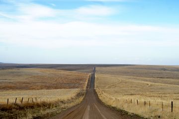 A long and hilly dirt road cuts through a field. The field is shades of harvest yellow and reddish-orangish-brown and has a barbed wire fence the length of the field. The sky is mostly white with a hint of blue at the top of the image.