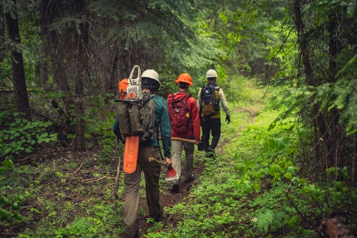 Three people walking along a mountain bike trail, surrounded by lush green forest. The volunteers are wearing hard hats and carrying trail stewardship tools, including a chainsaw.