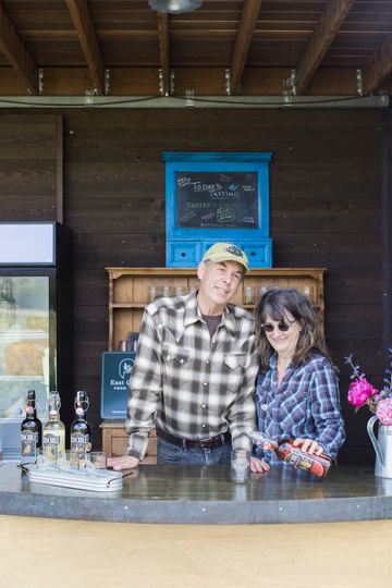 A couple poses behind the Runcible Cider tasting counter. A slightly obscured East Gorge Food Trail sign is visible behind them.