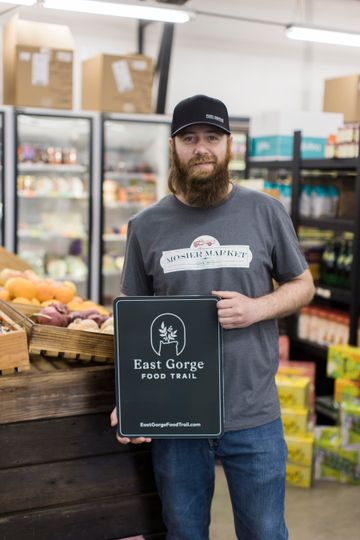 A man wearing a Mosier Market branded t-shirt poses with an East Gorge Food Trail sign.