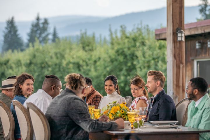 Ten people sit at long dining table with yellow flower centerpiece; vineyard and green hills in the background.