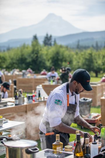 Man leans over outdoor cooking station in orchard setting, Mt. Hood in the background.