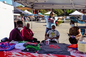 4 women sit at vendor table displaying t-shirts, 5thwoman stands among them with arms outstretched.